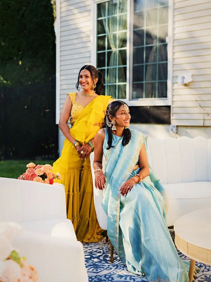 Mother and daughter in yellow and pastel blue South Asian attire on a white scalloped garden lounge with coral florals