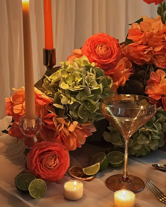 Coral ranunculus, orange taper candles, green hydrangea and limes around a coupe glass on a candlelit table