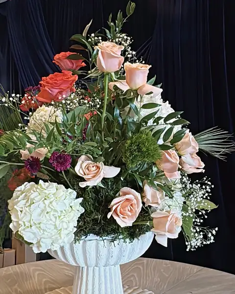 Fresh peach roses, white hydrangea, coral roses and baby’s breath in a white pedestal vase against navy velvet
