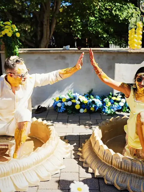 Bride and groom covered in turmeric giving a high-five from gold lotus haldi tubs beside blue and white florals