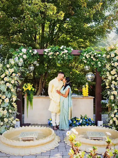 Couple embracing under a floral pergola of white roses, greenery and mirror balls with gold lotus haldi tubs in front