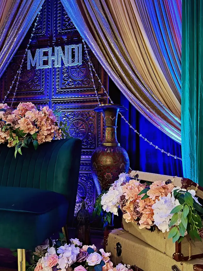 Mehndi lounge with a carved door backdrop, crystal MEHNDI sign, jewel-tone draping, emerald velvet sofa and peach florals