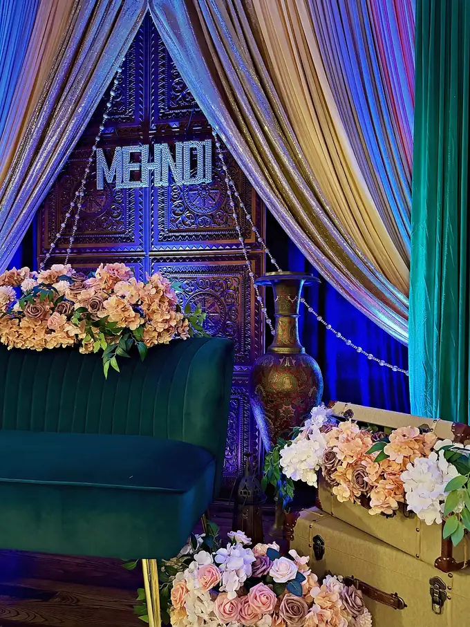 Mehndi lounge with a carved wooden door, crystal MEHNDI sign, jewel-tone draping, emerald velvet sofa and peach floral trunks