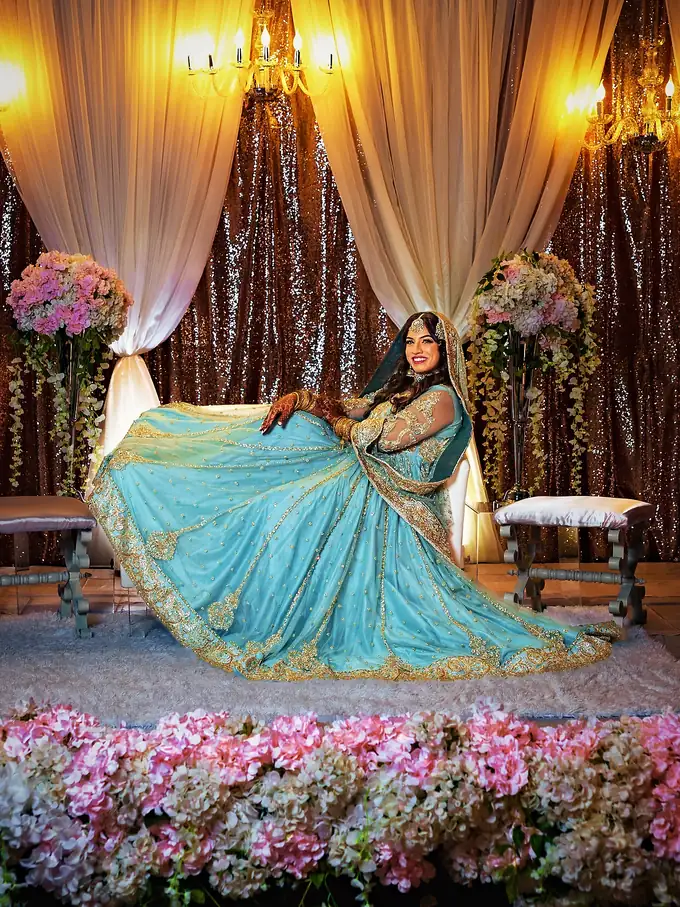 Bride in a sky blue lehenga on a rose gold sequin reception stage with ivory drapes, crystal sconces and blush florals, designed by Eden Events & Co.