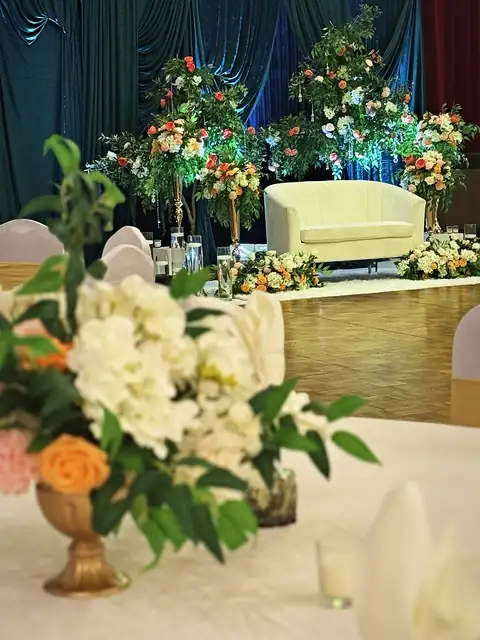 Ivory loveseat reception stage with towering garden rose arrangements and emerald velvet, seen past a centerpiece