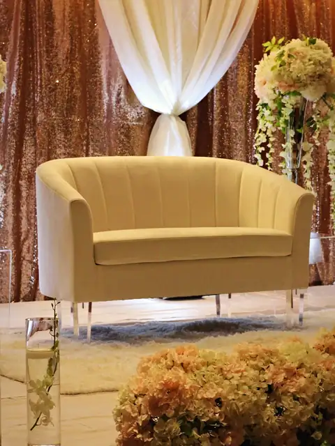 Ivory loveseat on a reception stage with rose gold sequin backdrop, ivory drapes and hanging floral stands