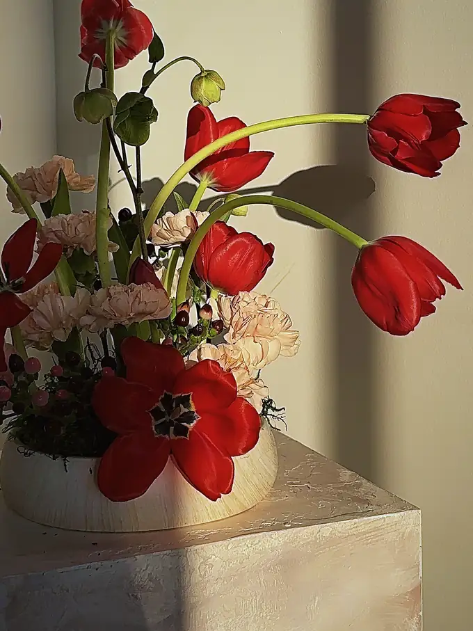 Red tulips and peach carnations arching from a ceramic bowl on a plaster plinth in golden studio light, by Eden Events & Co.