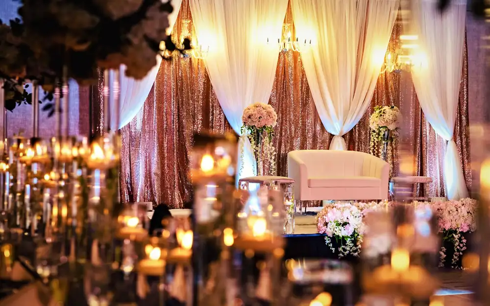 Rose gold sequin reception stage with ivory drapes, chandeliers and blush florals seen through glowing floating candles, by Eden Events & Co.