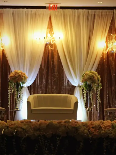 Reception stage with rose gold sequin backdrop, ivory drapes, crystal sconces, an ivory loveseat and a blush floral riser