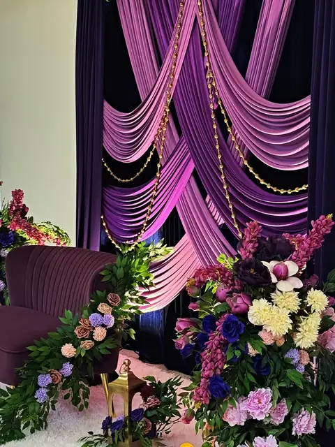 Crossed lilac drapes over plum velvet with gold bead strands, a plum loveseat and purple and blush floral columns