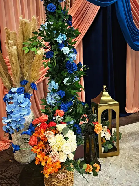 Sangeet backdrop of royal blue drapes swagged over peach, with a floral column of blue orchids, coral roses and pampas beside a gold lantern