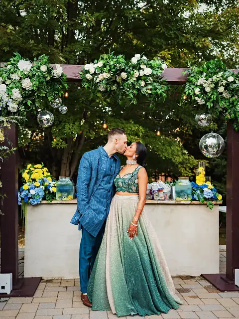 Couple in sangeet outfits kissing at a lemon bar framed by white roses, mirror balls and blue and yellow florals