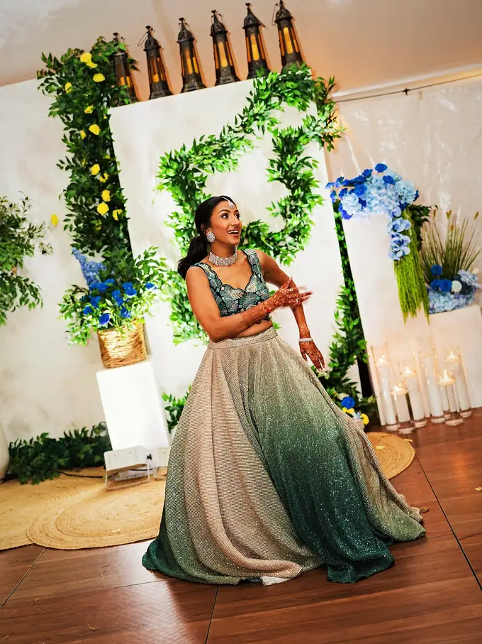 Bride dancing at her sangeet in front of a greenery monogram wall with gold lanterns, blue florals and candlelight, designed by Eden Events & Co.