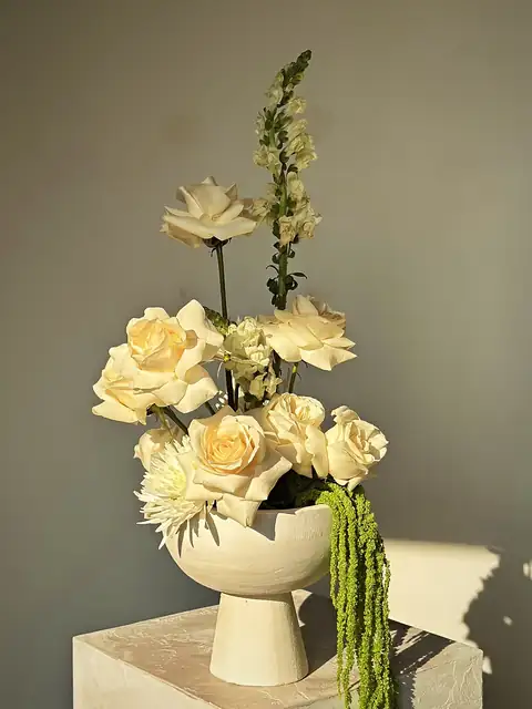 White roses and snapdragon in a sculpted white bowl with hanging amaranthus and a sun shadow